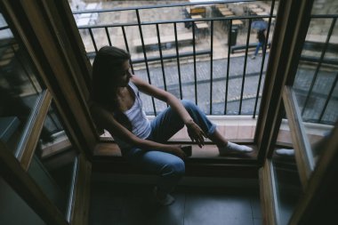 Pencerenin kenarında oturan genç bir kadın şık ve sakin bir şehir ortamında hayata kafa yoruyor.