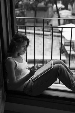 Kendini adamış bir sanatçı, doğanın güzelliğiyle çevrili, pencerenin kenarındaki rahat bir köşeye sakince resim çizer.