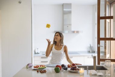 Neşeli bir kadın taze meyvelerle dolu parlak bir mutfakta ustalıkla peynirle hokkabazlık yapıyor.