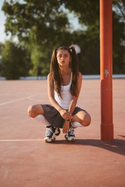 Genç bir kız, sıcak güneş ışığı altında kaygısız bir anın tadını çıkararak bir basketbol sahasında neşeyle çömeliyor.