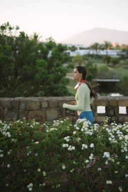 Genç bir kadın çiçeklerle ve yemyeşil çiçeklerle çevrili manzaralı bir yolda enerjik bir şekilde koşar.