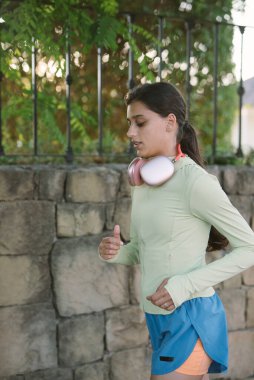 Kararlı bir kız güzel bir taş duvarın yanında koşuyor, kulaklıkla canlandırıcı egzersizinin tadını çıkarıyor.