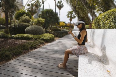 Genç bir kadın VR kulaklığıyla yeni sanal dünyaları keşfederken parkta huzurlu bir anın tadını çıkarıyor.