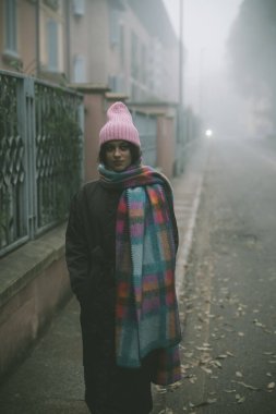 Pembe şapkalı ve yumuşak eşarplı bir kadın gizemli bir atmosferle çevrili sisli bir sokakta duruyor.