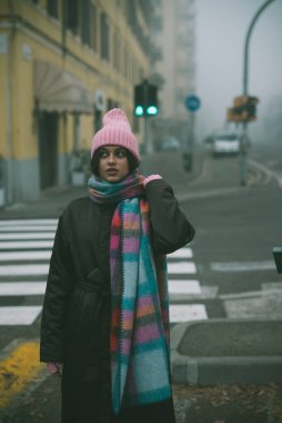 Pembe şapkalı ve eşarplı bir kadın sisli bir sokakta duruyor gizem ve entrikayla dolu.