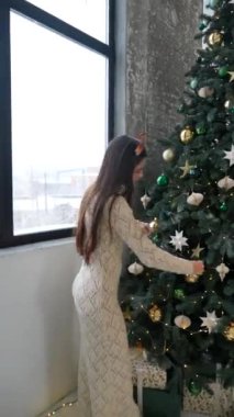 Bir kadın evin içinde güzel bir Noel ağacını dikkatlice süslüyor, rahat bir ortamda neşe ve tatil neşesi yayıyor.