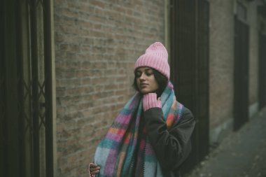 Bir kadın, sağlam tuğla bir duvarın önünde kendine güvenerek duruyor. Pembe bir şapka ve uyumlu pembe bir eşarpla süslenmiş.