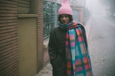 Şık pembe bir şapka ve rahat bir eşarp takan bir kadın sisli, sisli bir sokakta duruyor ve donuk bir çevreye renk katıyor.
