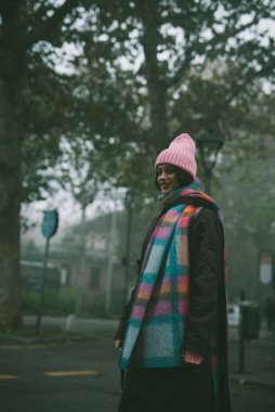 Parlak pembe bir şapka ve canlı, renkli bir eşarp ile şık bir şekilde süslenmiş bir kadın canlı bir sokakta dikiliyor.