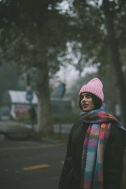 Canlı pembe bir şapka ve eşarpla süslenmiş bir kadın hareketli bir caddede zarif bir şekilde dikiliyor ve etrafını sarıyor.