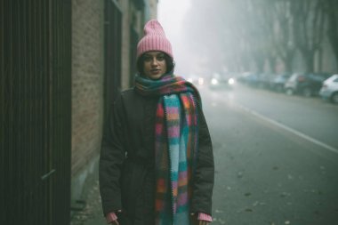 Pembe şapkalı ve eşarplı bir kadın sisli, sisli bir sokakta dikiliyor ve atmosferik bir manzara yaratıyor.
