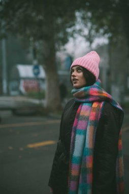 Meşgul bir caddede duran bir kadın şık pembe bir şapka takıyor ve göz kamaştırıcı, renkli bir eşarp giyip elbisesine cazibe katıyor.