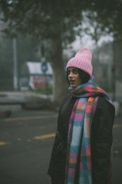 Meşgul bir caddede duran bir kadın şık pembe bir bere takıyor ve elbisesine renk katan canlı, renkli bir atkı takıyor.