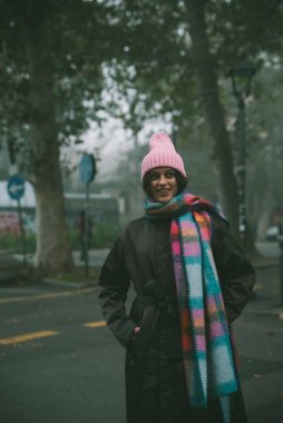 Şık pembe şapkalı ve eşarplı bir kadın şehrin hareketli caddesinde zarif bir şekilde duruyor.