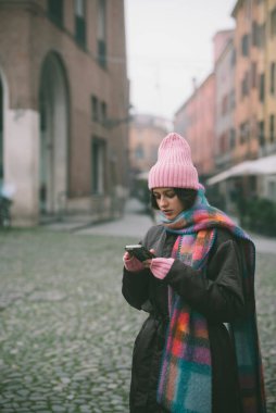 Şık pembe bir şapka takan ve eşarp takan bir kadın cep telefonuna bakıyor, belki de mesajlarını ya da sosyal medya hesaplarını kontrol ediyor.