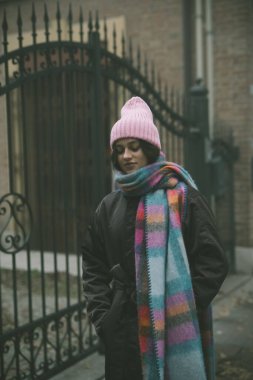 Zarif bir şekilde parlak pembe bir şapka ve güzel renkli bir eşarpla süslenmiş bir kadın dayanıklı ahşap bir çitin önünde duruyor.