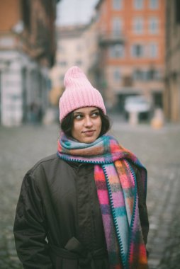 Parlak ve renkli bir eşarpla birlikte canlı pembe bir bere takan bir kadın büyük ve ilginç bir binanın önünde duruyor.