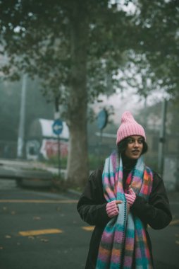 Şık pembe bir şapka ve eşarp takan bir kadın sokakta zarif bir şekilde duruyor ve sahneye renk katıyor.