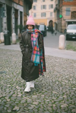 Şık pembe şapkalı ve rahat eşarplı bir kadın eski bir kaldırım taşı caddesinde yürüyor ve çevresinin tadını çıkarıyor.