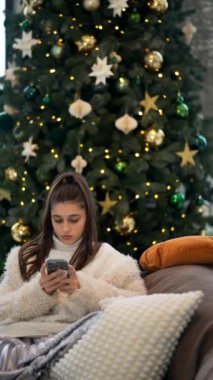 Genç bir kız, güzel dekore edilmiş bir Noel ağacıyla akıllı telefonuna odaklanmış bir şekilde koltukta dinleniyor..