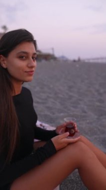 Sakin ve rahat bir kadın, huzurlu bir gün batımında elinde içkiyle güzel kumsalda eğleniyor.