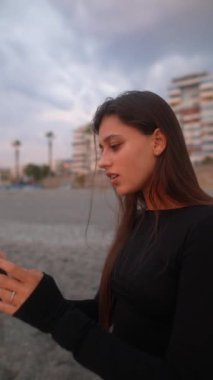 Şık siyah giyinmiş genç bir kadın gün batımında plajın keyfini çıkarırken akıllı telefonuyla meşgul oluyor.