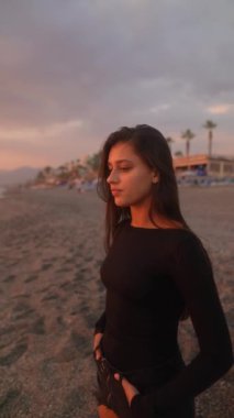 Genç bir kadın, güzel bir gün batımında, düşünceli bir şekilde uzaktaki sahile bakarak huzur içinde duruyor.