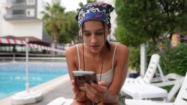 Güneşli bir havuz başında genç bir kadın, akıllı telefonuna dalmış, modern rahatlığı ve rahatlığı simgeliyor.