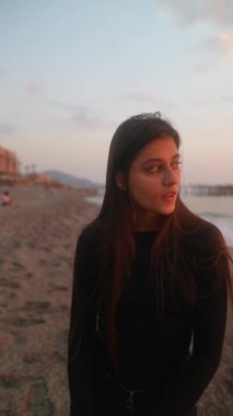 Genç bir kadın gün batımında plajda zarif bir şekilde duruyor, huzur ve sükunetin hislerini güzel bir şekilde yakalıyor.