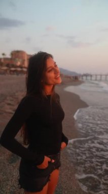 Genç bir kadın kıyıda durup, düşünceli bir şekilde güneşin batışını seyrediyor, bir huzur anı yakalıyor.