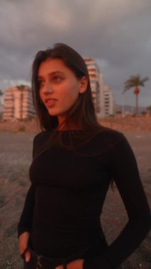 Zarif siyah elbiseler giymiş genç bir kadın gün batımında güzel bir sahilde düşünceli bir şekilde duruyor.