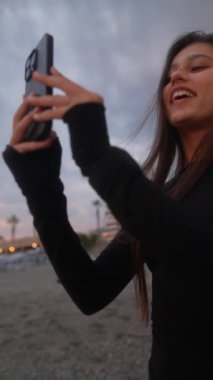 Şen şakrak bir gün batımı manzarası sırasında sahilde akıllı telefonuyla değerli anılarını yakalayan neşeli bir kadın.