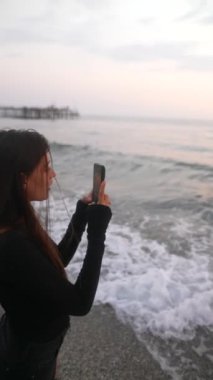 Genç bir kadın güzel bir kumsalda duruyor, akıllı telefonuna dalmış dalgalar yavaşça ayaklarına vuruyor.