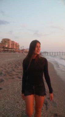 Genç bir kadın gün batımında sakin sahil şeridinde sakin bir sahil atmosferi yaratarak rahatça dolaşır.