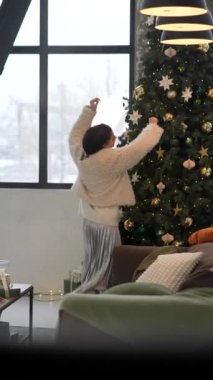 Bir insan, şık bir oturma odasında, dışarıda kış manzarası olan bir Noel ağacını yaratıcı bir şekilde süslüyor.