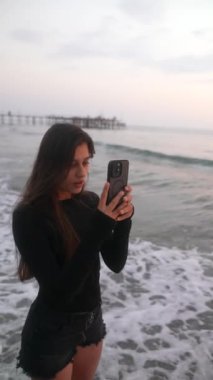 Genç bir kadın kumsalda güzel bir gün batımı ve arkasında hafif dalgalarla mutlu bir selfie çeker.