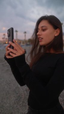 Bir kadın, güzel bir sahil günbatımının tadını çıkarırken akıllı telefonuyla değerli anları yakalamaktan zevk alır.