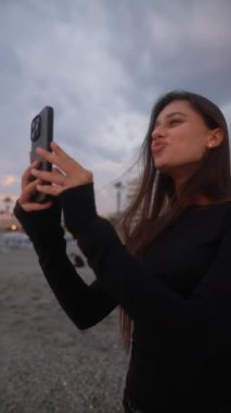 Neşeli genç bir kadın, etrafındaki büyüleyici deniz manzarasının tadını çıkarırken akıllı telefonunu mutlu bir şekilde kullanıyor.