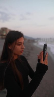 Genç bir kadın plajda büyüleyici günbatımı fotoğraflarını çekiyor, sanatsal yeteneğini ve iç huzurunu sergiliyor.