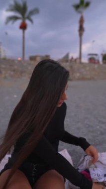 Sahilde neşeyle oturan genç bir kadının sakin bir sahnesi. Arka planda sallanan palmiyeler var.