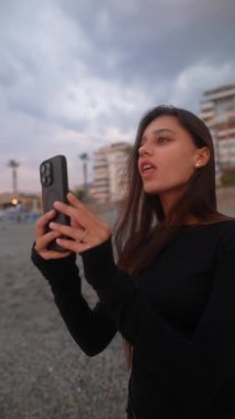 Genç bir kadın telefonuyla çarpıcı manzaralı bir kumsal zeminine karşı güzel anları yakalıyor, hayatın tadını çıkarıyor.