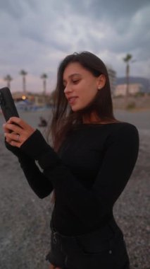 Neşeli bir kadın gün batımında güzel sahilde akıllı telefonunu kullanırken ışıl ışıl gülümsüyor.