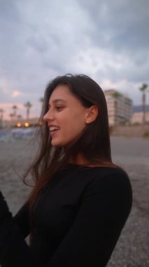 Neşeli bir kadın sahilde dikilirken heyecanlı bir şekilde selfie çeker arkasında palmiye ağaçları ve renkli bir gökyüzü vardır.