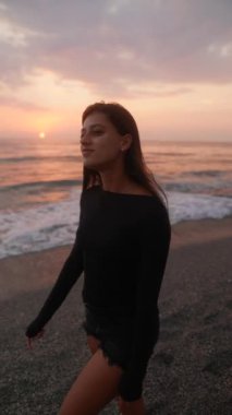 Genç bir kadın gün batımında güzel sahilde rahatça geziniyor, okyanus dalgalarının sükunetini kucaklıyor.