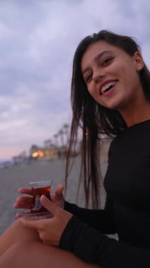 Bir kadın kumsalda dinlenmekten zevk alır. Sıcak çayını yudumlarken günbatımının huzurlu atmosferini seyreder.