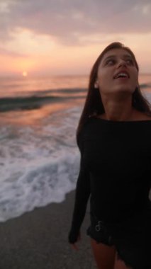 Güneş batarken, bir kadın sakin bir sahil boyunca sakin bir şekilde yürüyor ve güzel, huzurlu bir sahil atmosferi yaratıyor.