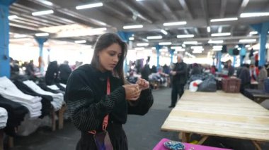 Genç bir kadın çeşitli kıyafetler ve canlı aksesuarlarla dolu hareketli bir markette rahatça dolaşır.