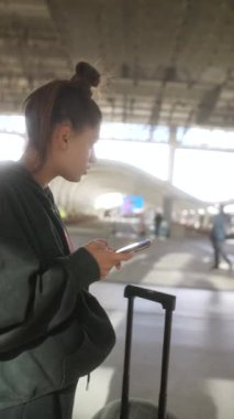 Yoğun bir havaalanındaki akıllı telefonuna dalmış genç bir kadın, bavuluyla birlikte seyahat titreşimlerini somutlaştırıyor.