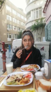 Bir kadın, güzel şehir mimarisi ve yemyeşil ile çevrili, rahat bir kafede lezzetli yemeğinin tadını çıkarıyor.