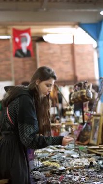 Genç bir kadın, antika eşyalar ve hazinelerle dolu hareketli bir pazarda çeşitli eşsiz eşyalar inceliyor.